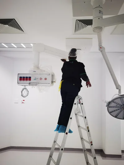 Cleaner on a ladder sanitising equipment arms in a hospital operating theatre